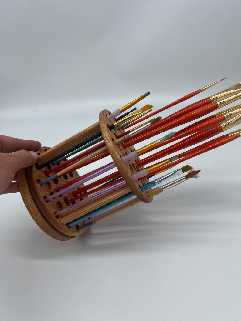 A wooden rotating paintbrush holder with a circular base and a cylindrical center. The holder has many holes to hold paintbrushes. The holder is filled with a variety of paintbrushes with different colored handles and bristles.
