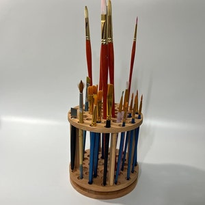 A wooden rotating paintbrush holder with a circular base and a cylindrical center. The holder has many holes to hold paintbrushes. The holder is filled with a variety of paintbrushes with different colored handles and bristles.