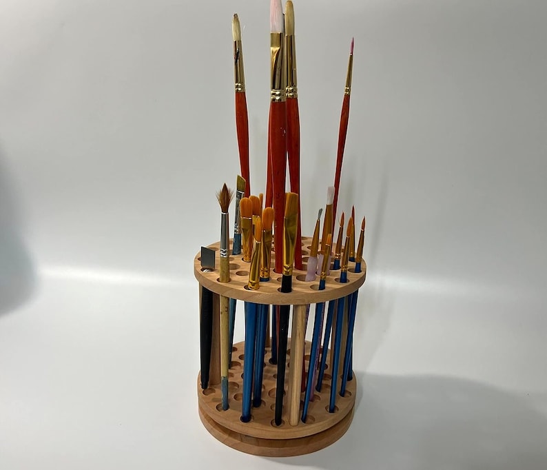 A wooden rotating paintbrush holder with a circular base and a cylindrical center. The holder has many holes to hold paintbrushes. The holder is filled with a variety of paintbrushes with different colored handles and bristles.