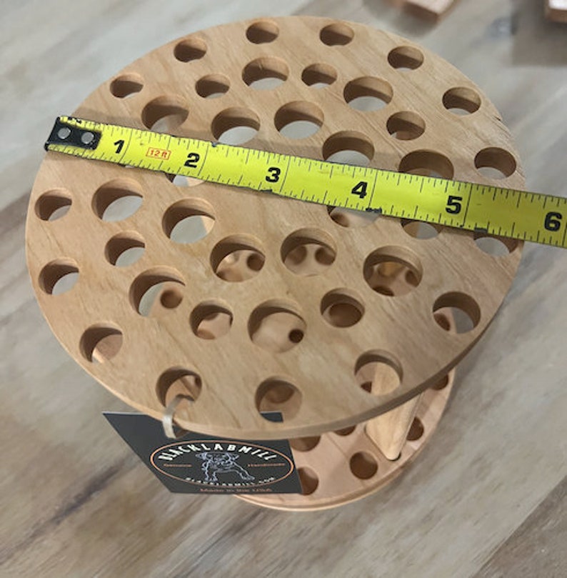 A wooden rotating paintbrush holder with a circular base and a cylindrical center. The holder has many holes to hold paintbrushes. The holder is filled with a variety of paintbrushes with different colored handles and bristles.