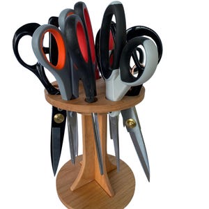 May include: A wooden scissor holder with a variety of scissors. The scissors have black, gray, and red handles. The holder is made of light-colored wood and has a round base.