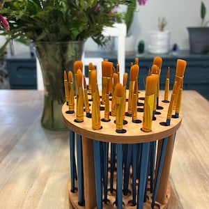 A wooden rotating paintbrush holder with a circular base and a cylindrical center. The holder has many holes to hold paintbrushes. The holder is filled with a variety of paintbrushes with different colored handles and bristles.