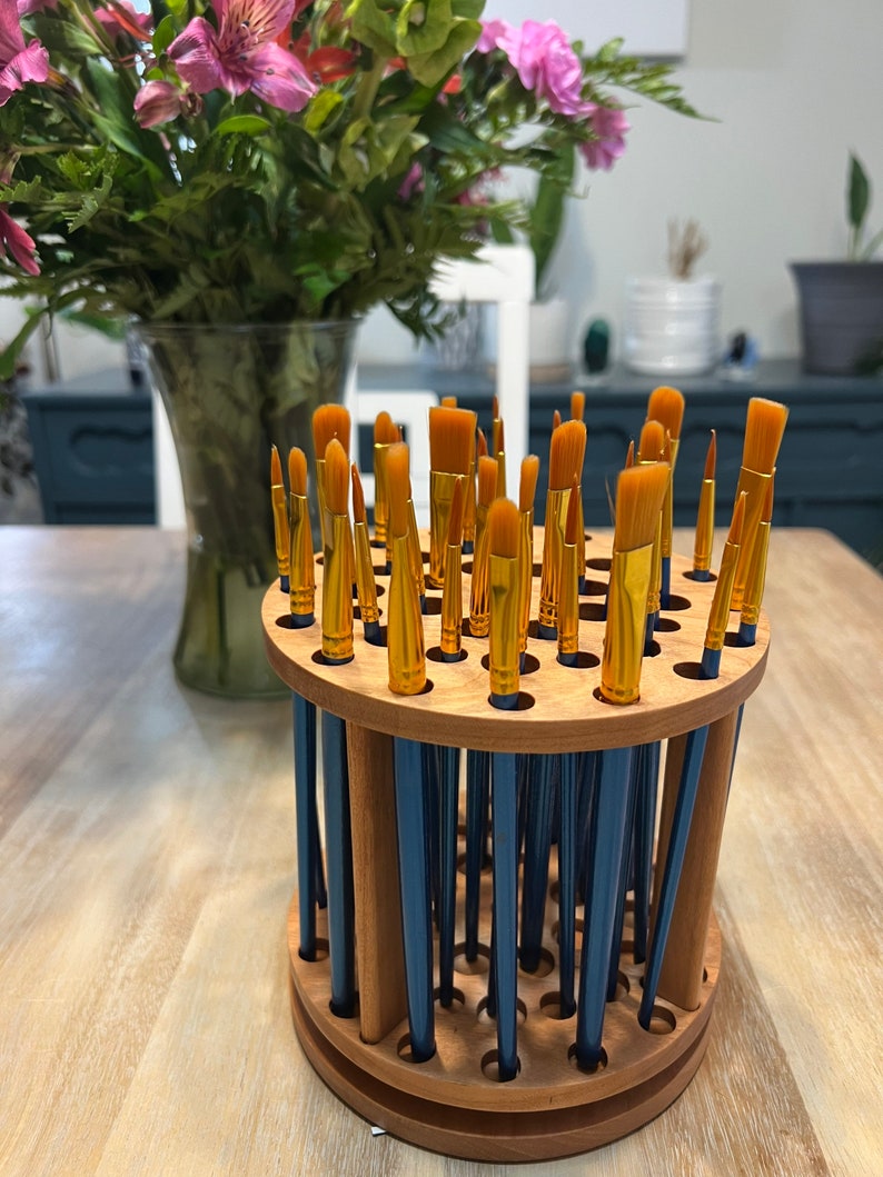 A wooden rotating paintbrush holder with a circular base and a cylindrical center. The holder has many holes to hold paintbrushes. The holder is filled with a variety of paintbrushes with different colored handles and bristles.