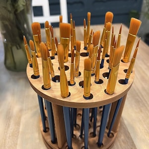 A wooden rotating paintbrush holder with a circular base and a cylindrical center. The holder has many holes to hold paintbrushes. The holder is filled with a variety of paintbrushes with different colored handles and bristles.