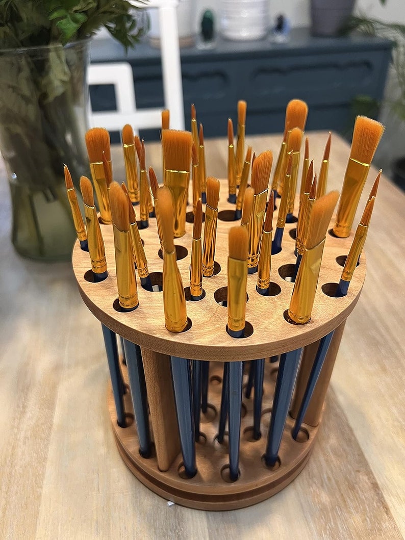 A wooden rotating paintbrush holder with a circular base and a cylindrical center. The holder has many holes to hold paintbrushes. The holder is filled with a variety of paintbrushes with different colored handles and bristles.