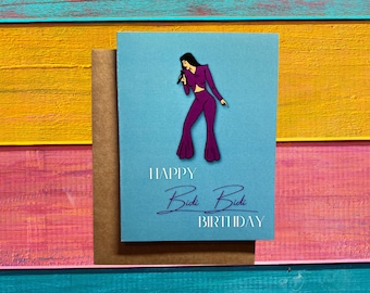 May include: A light blue birthday card with a purple illustration of a singer holding a microphone. The card reads "HAPPY Bidi Bidi BIRTHDAY" in white and purple script. The card is paired with a brown envelope.