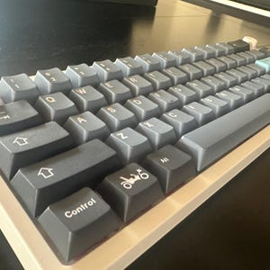 Könnte beinhalten: Eine weiße und graue mechanische Tastatur mit einer grauen Leertaste und einer blauen Taste. Die Tastatur hat einen weißen Rahmen und eine graue Tastenkappe mit einem kleinen Auto-Symbol.
