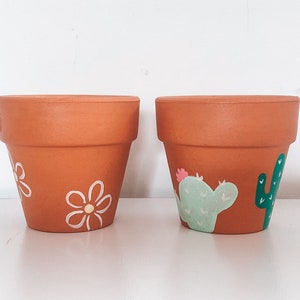 May include: Two terracotta flower pots, one decorated with white daisies and the other with green cacti and a pink flower.