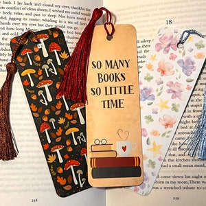 May include: Three colourful bookmarks with tassels. The first bookmark features a dark background with a pattern of red and orange mushrooms and leaves. The second bookmark has a tan background with the text "So Many Books So Little Time" and a stack of books with a coffee cup on top. The third bookmark has a white background with a watercolour pattern of pink, purple, and blue flowers.