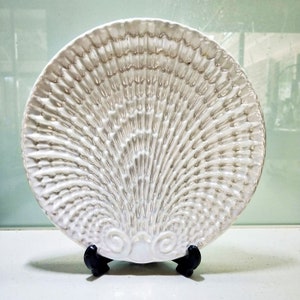May include: A white ceramic decorative plate shaped like a seashell with a textured surface. The plate is on a black stand.