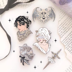 Peut inclure: Ensemble d'épingles en émail argenté avec des thèmes fantastiques. Les motifs comprennent une créature ressemblant à une chauve-souris, un buste avec des feuilles de laurier, une personne avec un bandeau, un casque de chevalier et une épée avec un papillon.