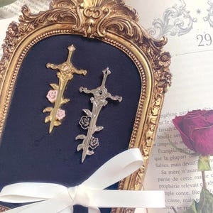 Può includere: Due spille a forma di spada in metallo su uno sfondo di velluto blu scuro all'interno di una cornice dorata decorata. Una spilla è color oro con accenti di rose rosa, l'altra è color argento con rose nere. Un nastro bianco è legato in basso.