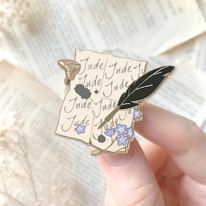 Peut inclure: Épingle en émail doré représentant un parchemin avec le mot "Jude" répété. Une plume noire et des myosotis bleus sont également inclus. L'épingle a un fermoir papillon.