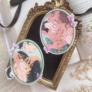 Pu&ograve; includere: Due adesivi ovali con illustrazioni di figure che si abbracciano, posti su uno specchio con cornice dorata. Un libro e fiori secchi sono visibili sullo sfondo. Sono presenti anche adesivi a farfalla rosa.