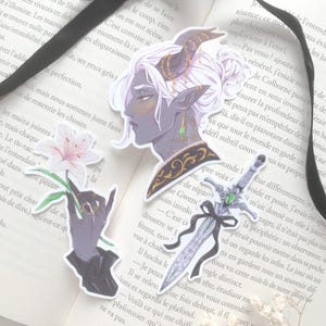 Może przedstawiać: Trzy naklejki na stronie książki: profil postaci z rogami, dłoń trzymająca kwiat i miecz z wstążką. Naklejki mają motyw fantasy z ciemną paletą kolorów.