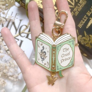 K&ouml;nnte beinhalten: Ein goldfarbener Schl&uuml;sselanh&auml;nger mit einem buchf&ouml;rmigen Anh&auml;nger mit dem Text "The Cruel Prince". Der Anh&auml;nger hat ein mintgr&uuml;nes und goldenes Farbschema mit einer Schlange und einem Schmetterlingsanh&auml;nger. Der Schl&uuml;sselanh&auml;nger wird in einer Hand gehalten.