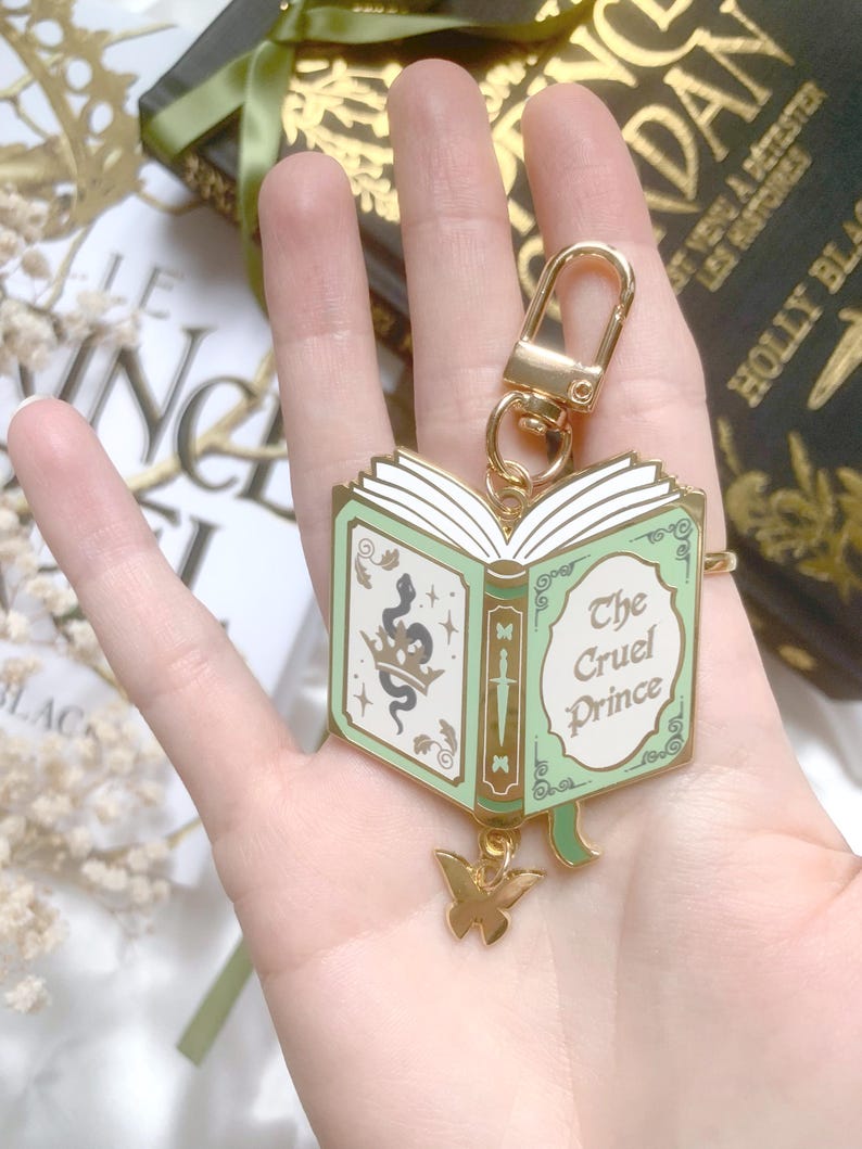 K&ouml;nnte beinhalten: Ein goldfarbener Schl&uuml;sselanh&auml;nger mit einem buchf&ouml;rmigen Anh&auml;nger mit dem Text "The Cruel Prince". Der Anh&auml;nger hat ein mintgr&uuml;nes und goldenes Farbschema mit einer Schlange und einem Schmetterlingsanh&auml;nger. Der Schl&uuml;sselanh&auml;nger wird in einer Hand gehalten.