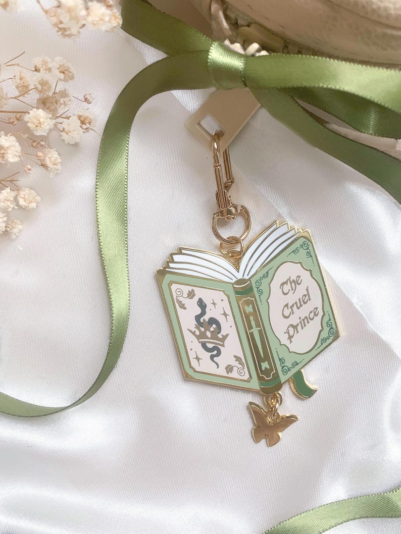 K&ouml;nnte beinhalten: Ein goldfarbener Schl&uuml;sselanh&auml;nger mit einem buchf&ouml;rmigen Anh&auml;nger mit dem Text "The Cruel Prince". Der Anh&auml;nger ist hellgr&uuml;n mit goldenen Akzenten und einem Schlangen- und Kronen-Design. Ein goldener Schmetterlingsanh&auml;nger h&auml;ngt darunter, und ein gr&uuml;nes Band ist oben gebunden.