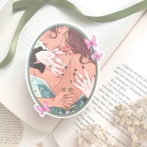Pu&ograve; includere: Un adesivo ovale con l'illustrazione di una persona con lunghi capelli castani, adornata da gioielli e con una spada. Farfalle rosa e un bordo verde incorniciano l'immagine, posta su un libro aperto.