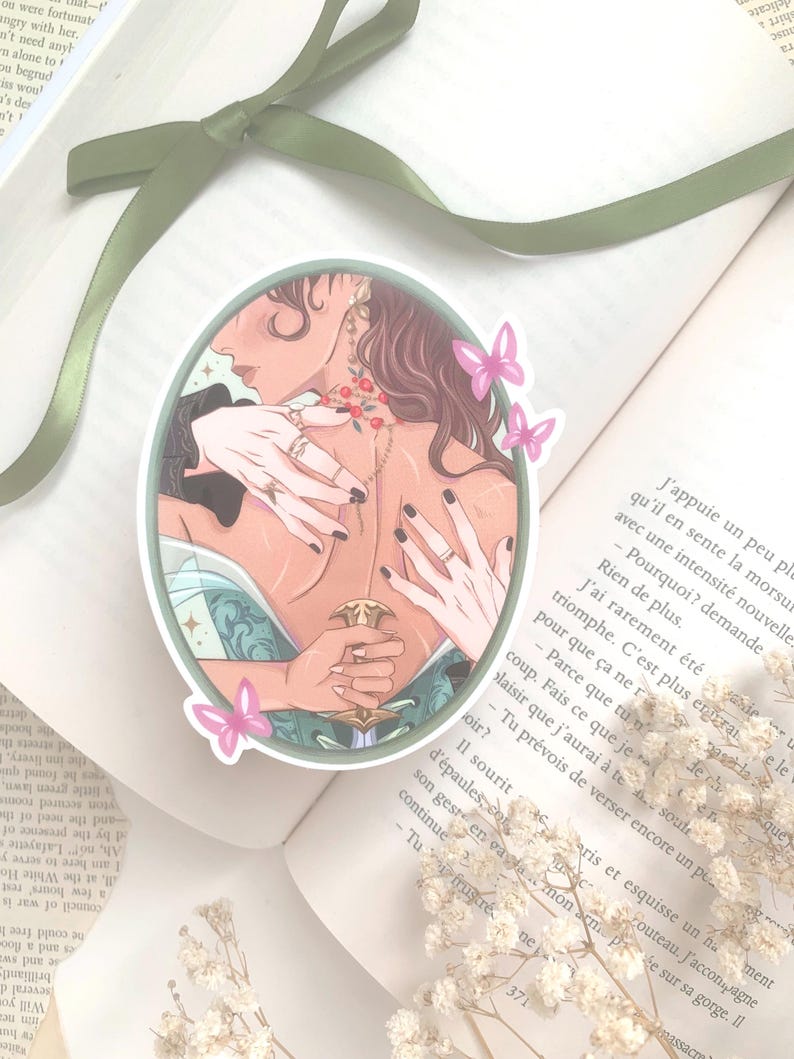 Pu&ograve; includere: Un adesivo ovale con l'illustrazione di una persona con lunghi capelli castani, adornata da gioielli e con una spada. Farfalle rosa e un bordo verde incorniciano l'immagine, posta su un libro aperto.