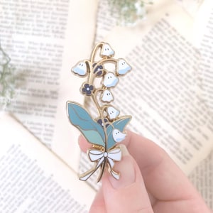 Op de afbeelding: Een goudkleurige emaille pin met een lelietje-van-dalen ontwerp. De pin heeft witte en lichtblauwe klokvormige bloemen, lichtblauwe bladeren en een witte strik. De pin wordt in een hand gehouden tegen een achtergrond van open boekpagina's.