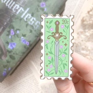 May include: A green and white enamel pin shaped like a postage stamp with a gold sword and flowers. The pin says "20c" in the top right corner.