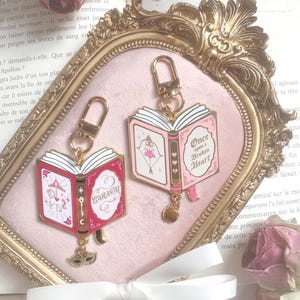 Peut inclure: Deux porte-clés en forme de livre ouvert avec des attaches dorées. L'un porte le mot "CARAVAL" sur la couverture, l'autre "Once Upon a Broken Heart". Les deux ont des breloques pendantes et des accents roses et rouges.