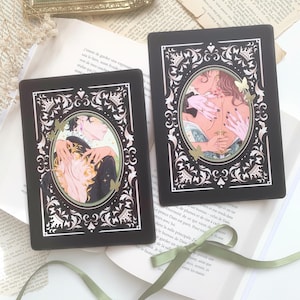 May include: Two black rectangular art prints with oval illustrations of romantic scenes. The illustrations feature figures in embrace, with soft colour palettes and decorative borders. The prints are on a background of open books and a green ribbon.