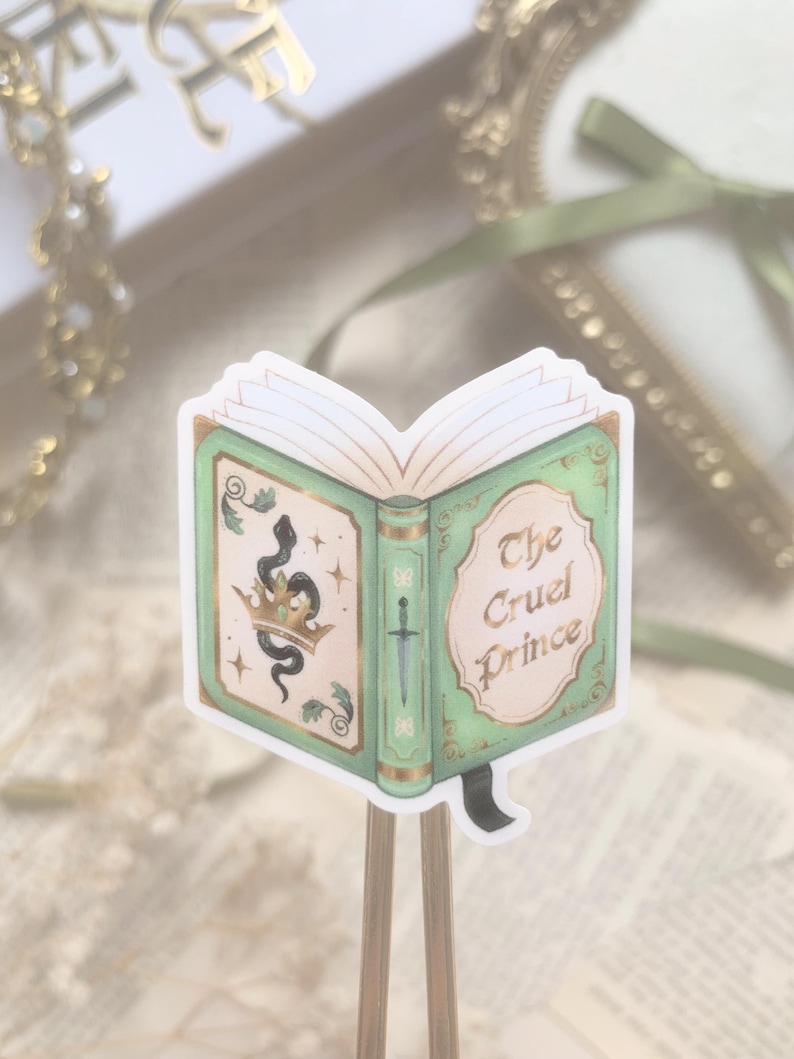 May include: A sticker of an open book with a green cover and gold accents. The book's cover features a snake, crown, and sword design. The title "The Cruel Prince" is written on the right side of the book.