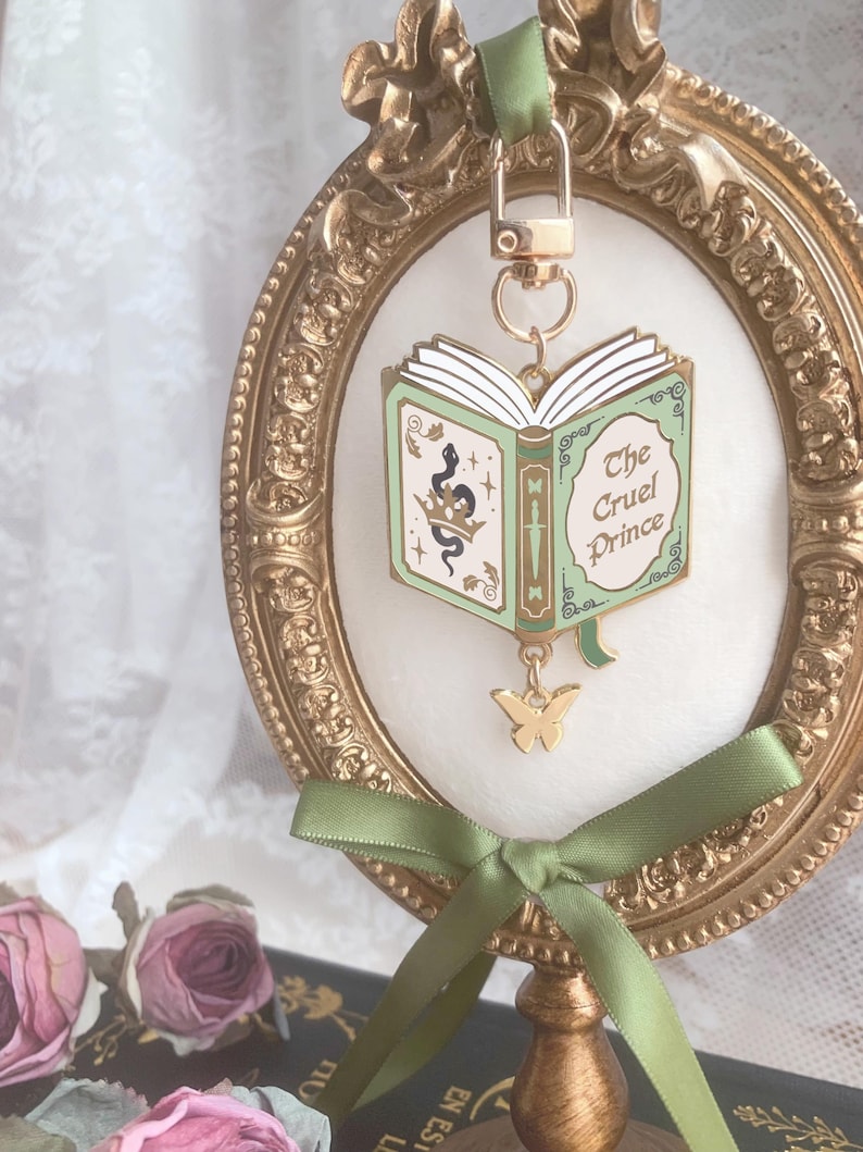 K&ouml;nnte beinhalten: Ein goldfarbener Metall-Schl&uuml;sselanh&auml;nger mit einem Buchdesign und dem Text "The Cruel Prince". Das Buch ist ge&ouml;ffnet, mit gr&uuml;nen Einb&auml;nden und wei&szlig;en Seiten. Ein goldener Schmetterlingsanh&auml;nger h&auml;ngt darunter. Der Schl&uuml;sselanh&auml;nger ist an einem gr&uuml;nen Band befestigt und in einem goldenen Rahmen ausgestellt.