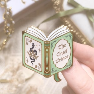 Pode incluir: Um alfinete de esmalte com bordas douradas em forma de livro aberto. O livro tem uma capa verde-menta com um desenho de cobra e coroa na página esquerda e as palavras "The Cruel Prince" na página direita.