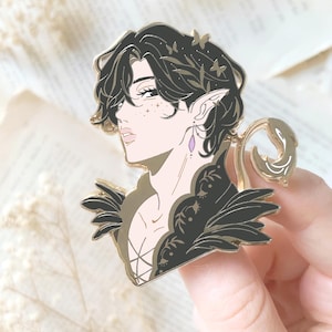 Op de afbeelding: Een goudkleurige emaille pin met een gestileerde illustratie van een persoon met donker haar, puntige oren en een decoratieve kraag. De pin heeft een vlinder- en bladerontwerp en een paarse oorbel.