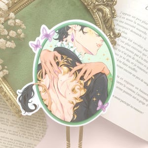 Pu&ograve; includere: Un adesivo decorativo con un'illustrazione di due figure che si abbracciano, all'interno di un cerchio verde. L'opera presenta una figura maschile con capelli scuri e un cappotto nero ornato da accenti dorati. Farfalle e stelle viola decorano il design.