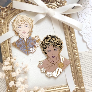 May include: Two enamel pins featuring stylised portraits of individuals. One pin depicts a blonde person in a blue patterned tunic with white flowers. The other pin shows a dark-haired person with a gold laurel wreath, wearing a white toga and gold jewellery.