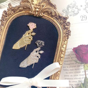 Può includere: Due spille in smalto metallico a forma di mani che tengono una rosa. Una spilla è color oro con una rosa rosa, l'altra è color argento con una rosa nera. Entrambe presentano linee dettagliate e un motivo testurizzato sul polso.