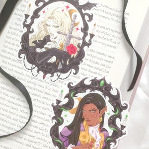 May include: Two illustrated stickers featuring characters from a fantasy novel. The first sticker features a blonde man with a sword and a red rose. The second sticker features a woman with long dark hair and a golden crown. Both stickers are framed in black with green accents.