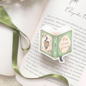 May include: A green and gold book-shaped sticker with the text "The Cruel Prince" on an open book. The sticker features a crown, snake, and sword design. A green ribbon and a gold crown are also visible.