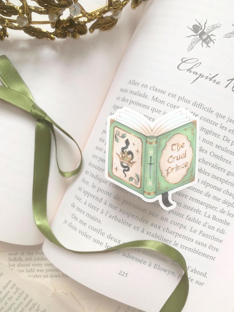 May include: A green and gold book-shaped sticker with the text "The Cruel Prince" on an open book. The sticker features a crown, snake, and sword design. A green ribbon and a gold crown are also visible.