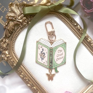 K&ouml;nnte beinhalten: Ein goldfarbener Schl&uuml;sselanh&auml;nger mit einem Buchdesign in Gr&uuml;n und Wei&szlig; mit dem Titel "The Cruel Prince". Er hat einen goldenen Schmetterlingsanh&auml;nger und einen goldenen Verschluss. Der Hintergrund umfasst einen goldgerahmten Spiegel, gr&uuml;nes Band und eine Rose.