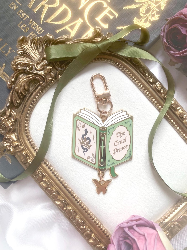 K&ouml;nnte beinhalten: Ein goldfarbener Schl&uuml;sselanh&auml;nger mit einem Buchdesign in Gr&uuml;n und Wei&szlig; mit dem Titel "The Cruel Prince". Er hat einen goldenen Schmetterlingsanh&auml;nger und einen goldenen Verschluss. Der Hintergrund umfasst einen goldgerahmten Spiegel, gr&uuml;nes Band und eine Rose.
