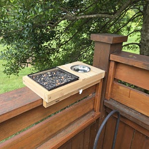 May include: A light-colored wooden bird feeder platform designed to mount onto a railing. It features a square black tray filled with mixed birdseed and a circular silver metal bowl embedded into the wood surface, secured by a white adjustment knob.
