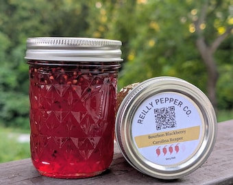 May include: A glass jar with a diamond-patterned texture filled with a deep red liquid containing dark seeds, topped with a silver lid. Beside it, a second silver lid features a label reading REILLY PEPPER CO. Bourbon Blackberry Carolina Reaper.
