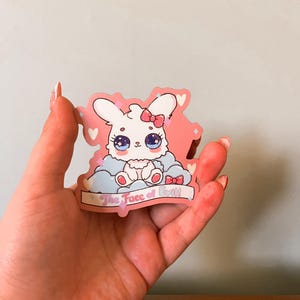 Peut inclure: Un autocollant représentant un lapin de dessin animé avec de grands yeux bleus, un nœud rose et un corps blanc duveteux. Le lapin est assis sur un nuage bleu avec une bannière indiquant "The Face of Doll". L'autocollant a une bordure rose avec des cœurs et des étoiles.