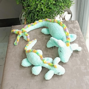 May include: Two crocheted plush toys in the shape of a sea creature, in a light blue color. The toys have a rainbow-colored spine and fins. The toys are lying on a neutral-colored surface.