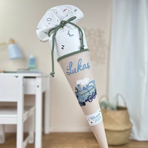 Könnte beinhalten: Ein beige-weißer Schultüte mit einem blauen Zugmotiv. Die Tüte ist mit dem Namen "Lukas" und dem Text "1. Klasse 2029" verziert.