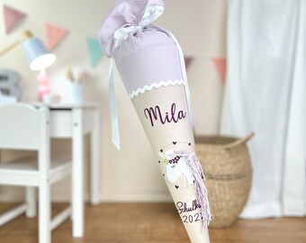 May include: A cone-shaped school gift bag made of light beige fabric with a lavender muslin top tied with a white ribbon. It features the name Mila, a unicorn illustration, and the text Schulkind 2024 in dark purple lettering.