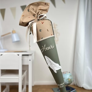 Könnte beinhalten: Ein grüner Schultüte mit einem weißen Walmotiv und dem Namen Henri, der auf die Seite gestickt ist. Die Tüte ist mit braunem Papier mit einem Muster aus bunten Buchstaben verziert.