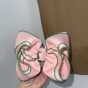 May include: A large, pink hair bow with green stitching along the edges. The bow is made of a satin-like material and is held in a person's hand. The background is a brown paper bag.