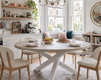May include: A round light wood dining table with a white X-shaped base, surrounded by four chairs with light wood frames and cream-colored upholstered seats. The table is set with white plates, clear glasses, and a bowl of fruit in the center.