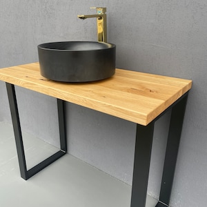 Può includere: Un lavabo in ceramica nero con un rubinetto dorato su un piano in legno con gambe in metallo nero.